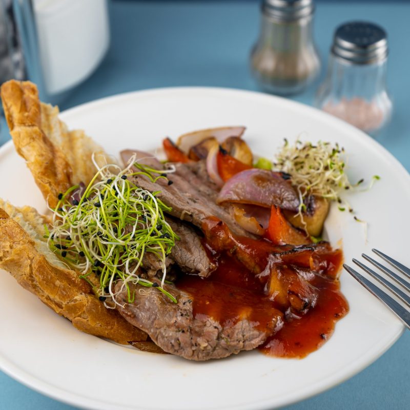 Warm salad with beef, vegetables, croutons and microgreen in a white plate on the restaurant table. Meat food in a bowl. Chop, medallions, cutlets. Blue background, close-up