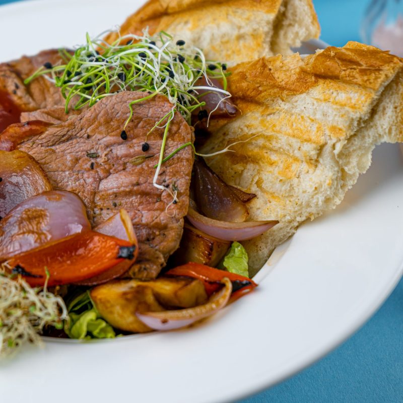 Warm salad with beef, vegetables, croutons and microgreen in a white plate on the restaurant table. Meat food in a bowl. Chop, medallions, cutlets. Blue background, close-up